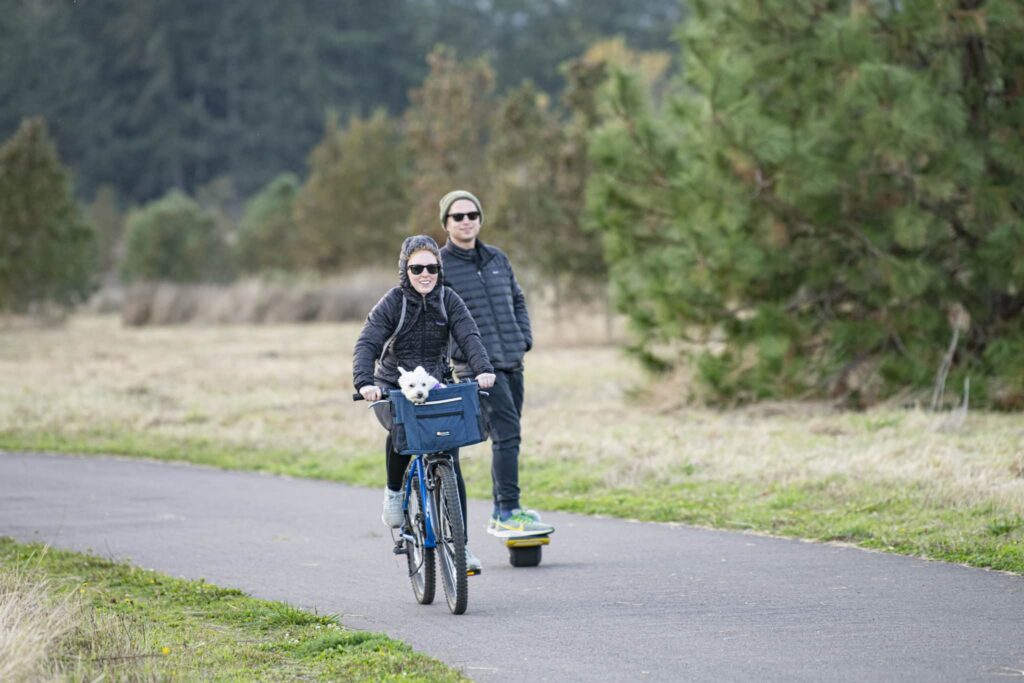 A woman biking with her dog in a basket and a man on a one-wheel