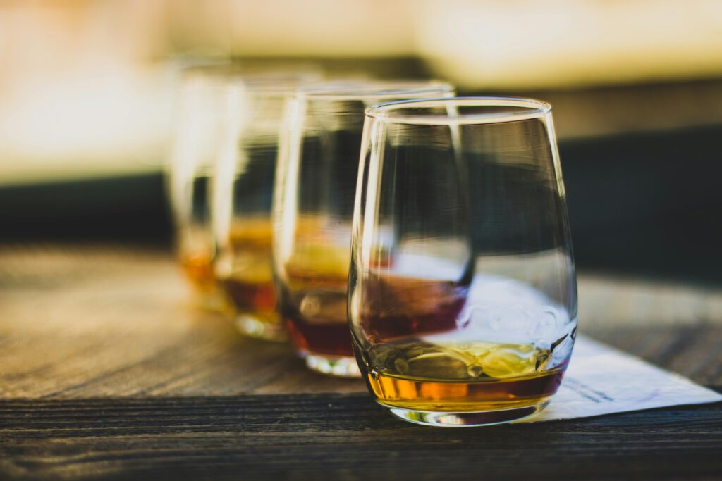 Photo of a flight of whiskey on a table. 4 small tasting glasses filled with a small amount of whiskey.