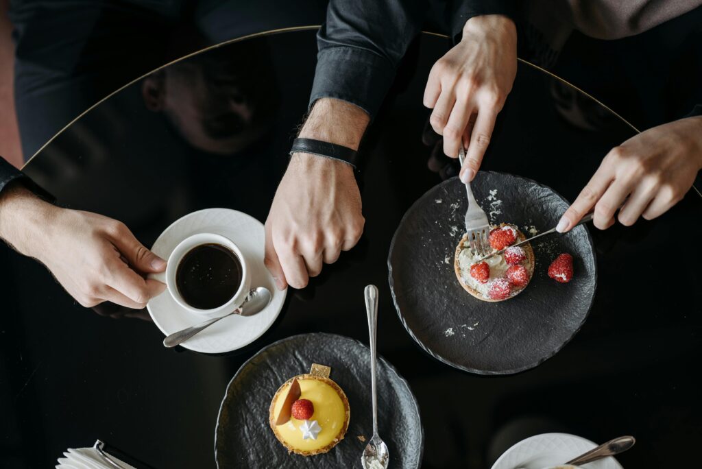 Photo of a pair of hands cutting a breakfast pastry along another pair of hands that are picking up a cup of black coffee. Photo is an aerial view of a table with two people sitting on one side of it.