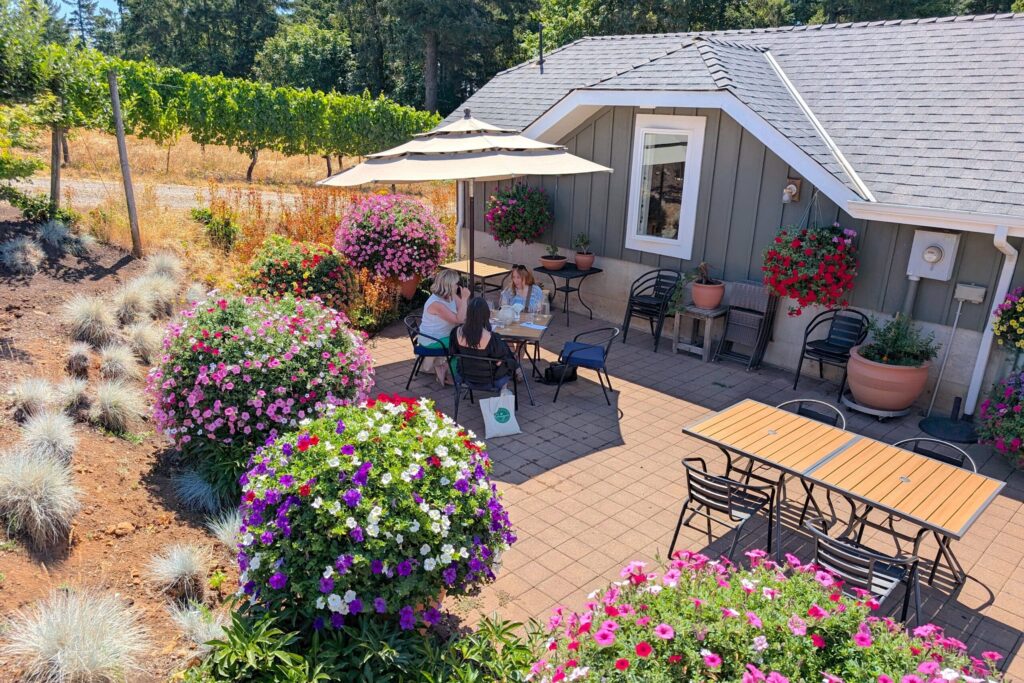 Photo of people enjoying the outdoor patio at Beckham Estate Vineyard surrounded by grape vines and flowers with the tasting room building in the background
